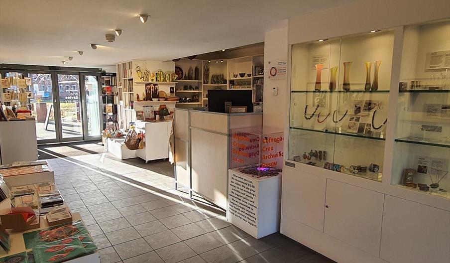 gallery shop inside view toward an entrance door. There are glass shelves on the right with jewelry inside and white plinths with books and cards of t